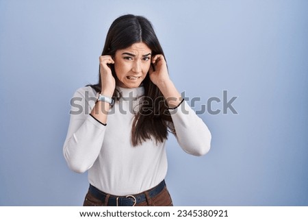 Young brunette woman standing over blue background covering ears with fingers with annoyed expression for the noise of loud music. deaf concept.  Royalty-Free Stock Photo #2345380921