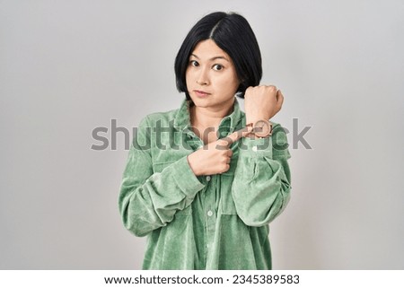 Young asian woman standing over white background in hurry pointing to watch time, impatience, looking at the camera with relaxed expression  Royalty-Free Stock Photo #2345389583