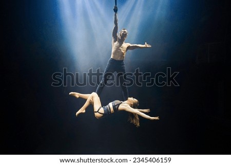 An acrobatic performance featuring an aerial straps duet: a man gracefully hangs by one hand, while a woman lies extended at his feet. They are bathed in an ethereal blue and white glow Royalty-Free Stock Photo #2345406159