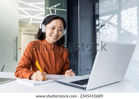 Asian business woman with headphones watching online training course at workplace, woman writing information happy and satisfied with the results of professional development