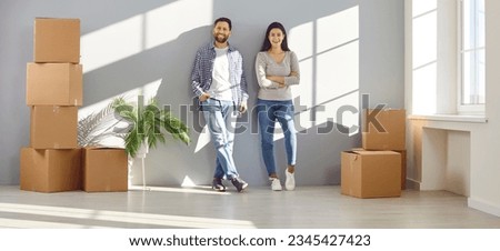 Happy couple standing against gray wall in empty room full of cardboard boxes. Happy homeowners standing in living room in their new apartment. Home relocation, family housing, real estate Royalty-Free Stock Photo #2345427423