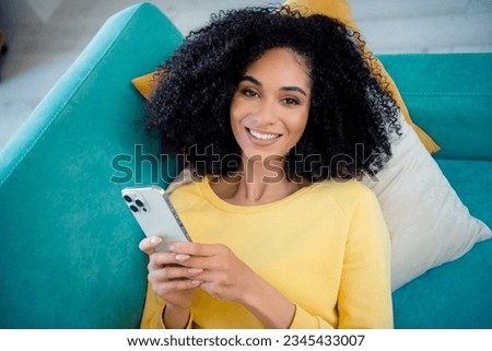 Close up photo of funny satisfied woman with perming coiffure wear yellow long sleeve lay on sofa hold smartphone at home room indoors