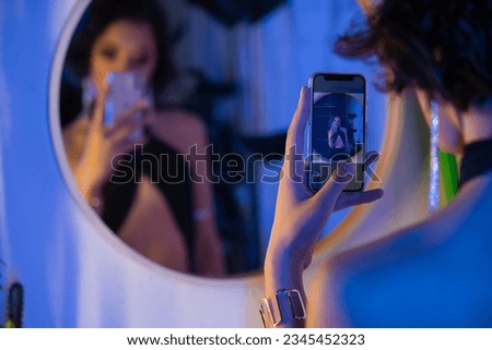 Blurred young asian woman in dress taking selfie on smartphone near mirror in night club