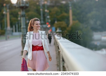 happy trendy middle aged woman in pink dress and white jacket in the city with red bag on the bridge. Royalty-Free Stock Photo #2345474103