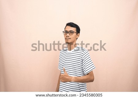 Asian man in glasses wearing casual striped shirt, posing giving thumbs up gesture or okay that agree. Isolated beige background.