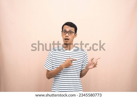 Asian man with glasses wearing casual striped shirt, whispering pose while pointing to the side. Isolated beige background.