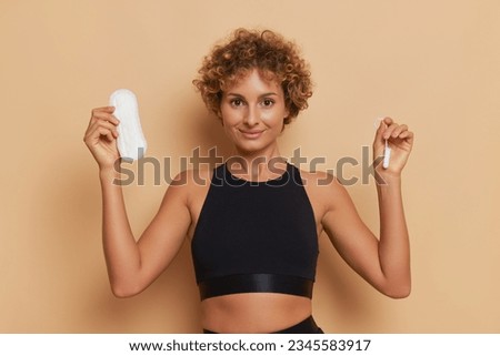 Curly woman in black top stands smiling with sanitary pad in hand, picture setup on light brown background, body hygiene concept, copy space