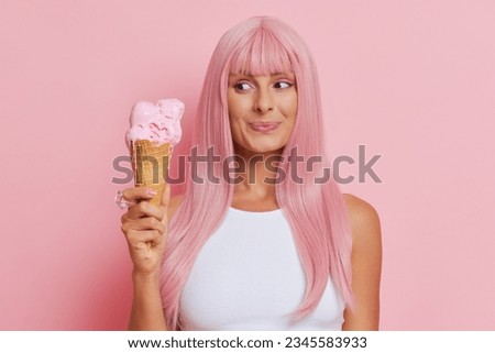 Beautiful girl in pink wig looks at ice cream in her hand with satisfied smile, picture setup on pink background, tasty food concept, copy space