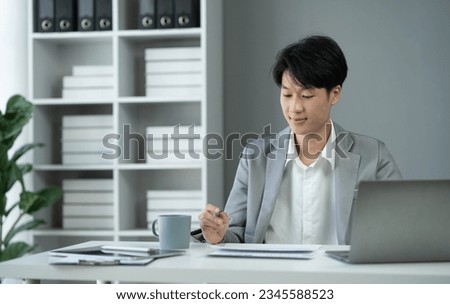 Asian young businessman sitting happily working in the office, calculating, analyzing, finding financial reports. Organize documents using laptop as a helper. Accounting, tax, management concept.