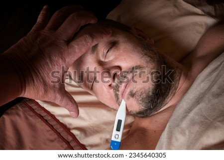 A young man is taking fever-reducing pills to lower his temperature. He is taking his body temperature to monitor his illness. A young man is feeling unwell and stays in bed.