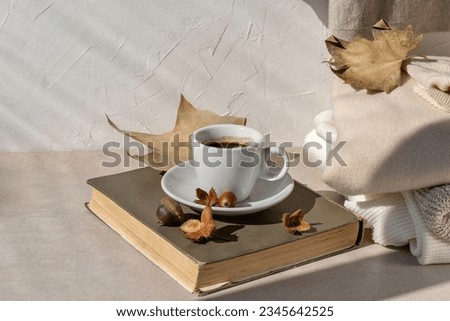 Hot coffee cup, book, fall light brown leaves, knitted sweaters in neutral colors on a beige table and white wall background with sunlight shadow. Aesthetic pastel autumn still life. Royalty-Free Stock Photo #2345642525