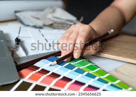 Woman picking paint colors from color scheme palette guide catalog with colour swatches. Design studio desk scene, creative work tools. Female architect or interior designer hand holding pencil.