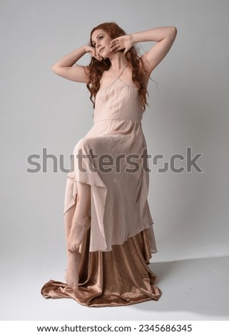 Full length portrait of beautiful  female model with long  brunette hair wearing a creamy pink gown dress. graceful sitting pose isolated on white studio background.