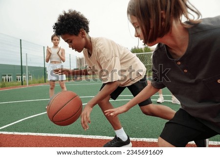 African American schoolboy in activewear dribbling ball while running forwards in front of his classmate while blond girl standing on background Royalty-Free Stock Photo #2345691609