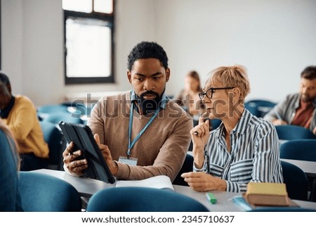 Senior woman and her African American professors using digital tablet during adult education training class in lecture hall.  Royalty-Free Stock Photo #2345710637