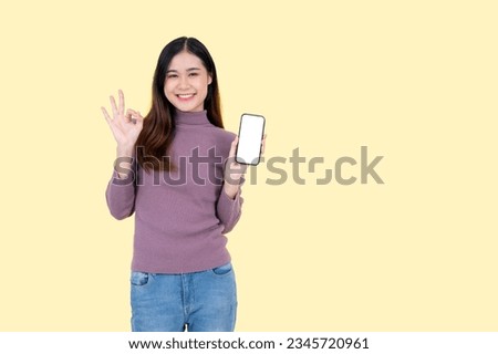A beautiful and smiling Asian woman in casual clothes shows the Okay hand sign, holds a smartphone mockup to the camera and stands against an isolated yellow background.