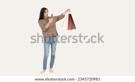 A cheerful and happy Asian woman is taking pictures of her shopping bags while standing against an isolated white background. shopping and lifestyle concept
