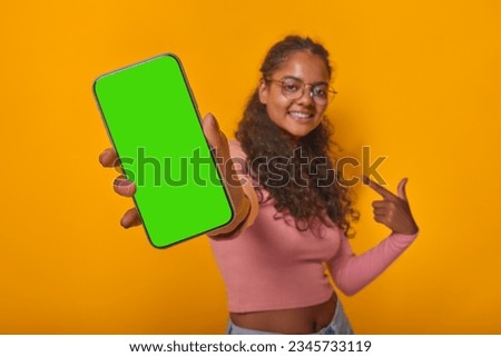 Young Indian woman holding phone with green screen and pointing finger at gadget smiling and offering to advertise application or website stands on isolated orange background. Copy space