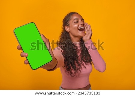 Young cheerful expressive Indian woman teenager with phone in hands shouts loudly announcing release of new mobile application or website for students stands in orange studio. Green screen