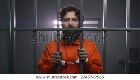 Prisoner in orange uniform holds metal bars, stands in prison cell in handcuffs, looks at camera. Criminal serves imprisonment term for crime in jail. Depressed inmate in detention center. Portrait. Royalty-Free Stock Photo #2345749565