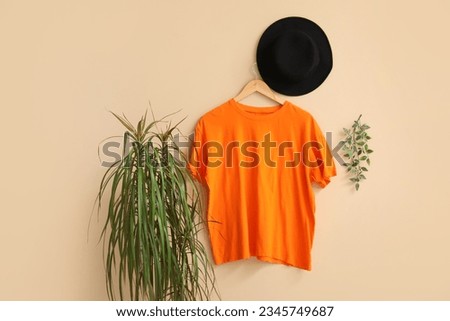 Stylish orange t-shirt and hat hanging on beige background