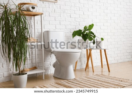 Interior of light restroom with ceramic toilet bowl, shelving unit and houseplants near white brick wall Royalty-Free Stock Photo #2345749915
