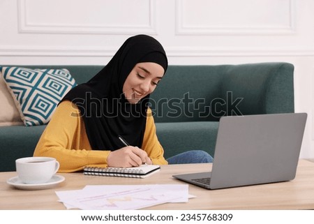 Muslim woman in hijab writing notes near laptop at wooden table indoors