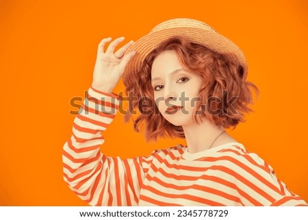 Beautiful bright girl with curly red hair, dressed in a straw hat and longsleeve with orange stripes posing on a yellow studio background. People's emotions. Summer fashion and beauty.