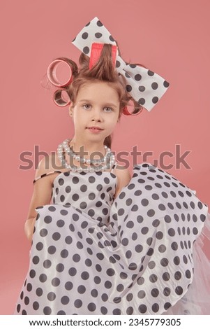 Fashion for kids - clothes and accessories. A little pretty girl in an elegant polka-dot dress and curlers on her head is going to a party. Pink studio background. Pin-up style.