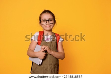 Advertising studio portrait of Caucasian chappy smiling elementary age school girl with backpack, holding a textbook, dressed in casual clothes and eyeglasses, looking at camera, isolated background Royalty-Free Stock Photo #2345818587