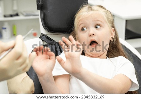 Childish fear. Cute little girl sitting in the dentist chair and covering her mouth in fear, being afraid of a dental drill in the hands of her doctor Royalty-Free Stock Photo #2345832169