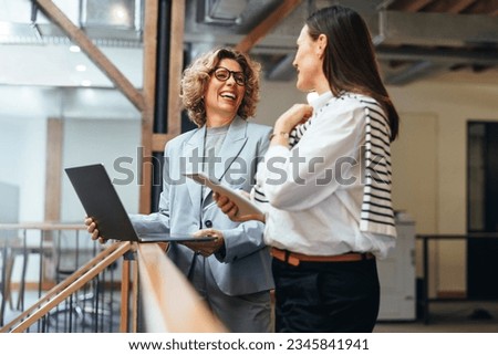Business women smiling as they have a meeting on a balcony. Professional business women discuss their upcoming work and ways to get it done. Women working together in a female-led startup. Royalty-Free Stock Photo #2345841941