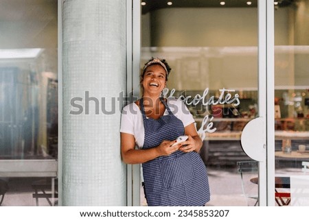 Successful cafe owner, a young woman, stands in her restaurant using a smartphone. Happy female entrepreneur using technology to manage her small business efficiently.