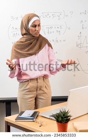 Vertical photo of puzzled Muslim female teacher leading online lesson. Tutor stands against the background of Mathematical board, looks at the laptop, shrugging his hands. Difficulty in learning