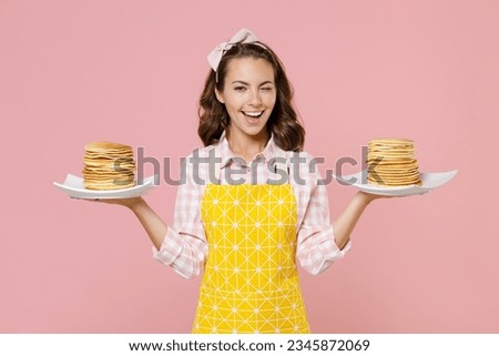 Blinking funny young brunette woman housewife wearing yellow apron hold plates with pancakes while doing housework isolated on pastel pink colour background studio portrait. Housekeeping concept Royalty-Free Stock Photo #2345872069