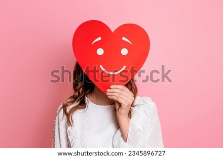 girl holding paper heart with smiley happy face isolated on pink background. Falling in love concept
