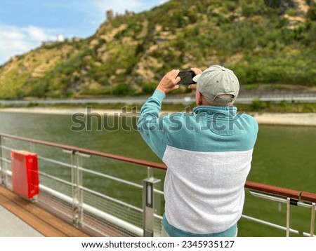 Rear view of a mature man using his mobile phone to capture a picture of the beautiful landscape. He is standing on the deck of a boat cruising on the Rhine River.  