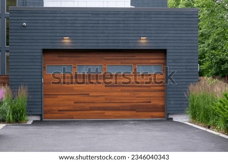 A modern brown faux wooden exterior garage door with four small horizontal glass windows. The modern door is on a luxury dark grey contemporary house with a concrete driveway.  Royalty-Free Stock Photo #2346040343