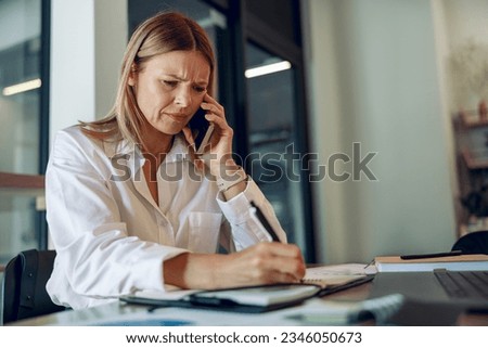 Worried woman entrepreneur making notes and talking by phone with client while sitting in coworking  Royalty-Free Stock Photo #2346050673