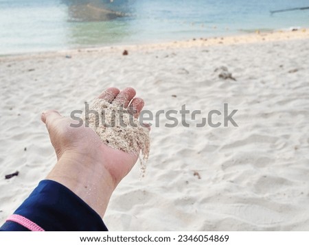 Fine sand in the hands of a woman. The left hand of tourist is pouring white sand on the ground while rest on a beach holiday. The background is sandy beach and sea with slight waves. Royalty-Free Stock Photo #2346054869
