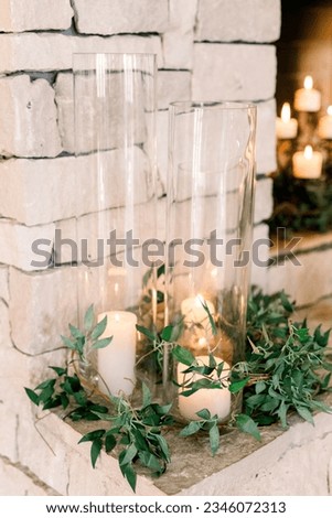 Elegant glass hurricane candle holders surrounded by greenery in front of a fireplace at a wedding reception. Royalty-Free Stock Photo #2346072313