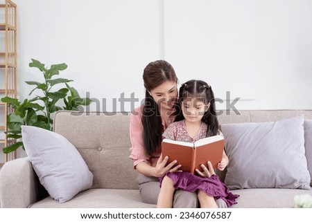 Attractive young asian woman nanny cuddling little kid, reading paper book together on comfortable sofa in living room. Happy mother educating small child daughter, enjoying weekend time at home.