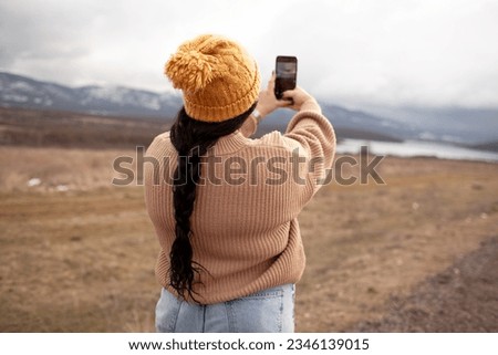 back view overweight model makes photo on phone. Beautiful woman is wearing jeans, sweater and hat walking in cold weather along the road  against the background of landscape and nature. 