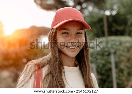 Portrait of beautiful teenage girl (14-15 years old) wearing a pink baseball cap, stands outdoors, sweetly smiling against the backdrop of a sunset. Royalty-Free Stock Photo #2346192127