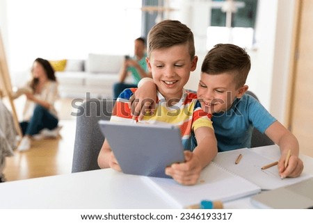 Two happy children watch an online lesson. Child uses tablet for learning and communication, and his younger brother is hugging him. Parents are sitting in background.