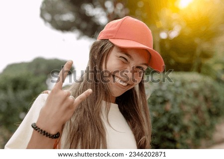 A vibrant smiling teenage girl winks, flashing a playful horn sign with one hand, reflecting the spirit of youth. She wears a pink baseball cap and looks trendy. Youth life and culture concept. Royalty-Free Stock Photo #2346202871