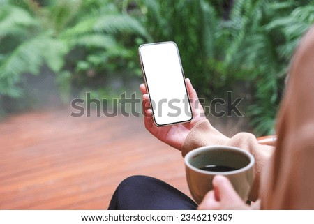 Mockup image of a woman holding mobile phone with blank white desktop screen while drinking coffee in the garden