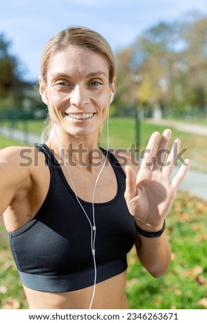 Female athlete after jogging in stadium talking with friends and recording online video blog, mature blonde with headphones looking at smartphone camera smiling, video call with headphones.