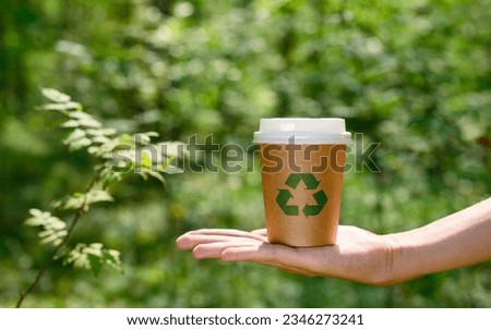 a paper cup with a recycling sign in hand against the backdrop of a forest. The concept of recycling and nature protection