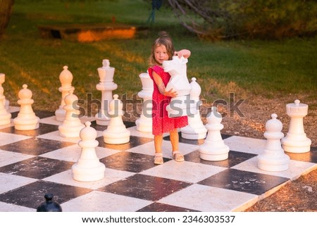 Little Caucasian girl in a pink dress playing street chess. A child plays with big chess pieces in the park on a summer evening. Royalty-Free Stock Photo #2346303537